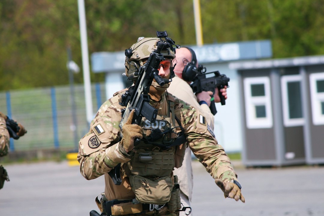 Operator w mundurze polowym, hełmie z mocowaniem noktowizora i zestawie łączności przemieszcza się z karabinkiem podczas ćwiczeń, w tle drugi operator osłaniający kierunek