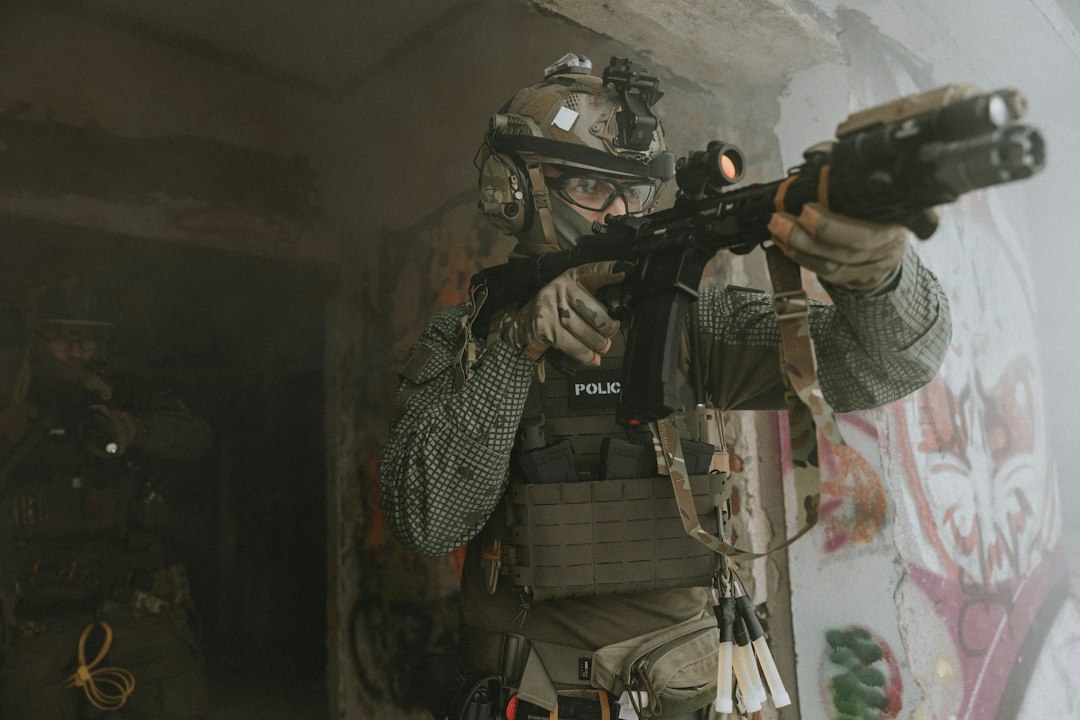 Operator w hełmie, okularach ochronnych i ochronnikach słuchu, z naszywką POLICE na kamizelce, celuje z karabinka wewnątrz zadymionego budynku, za nim w ciemnym przejściu drugi operator