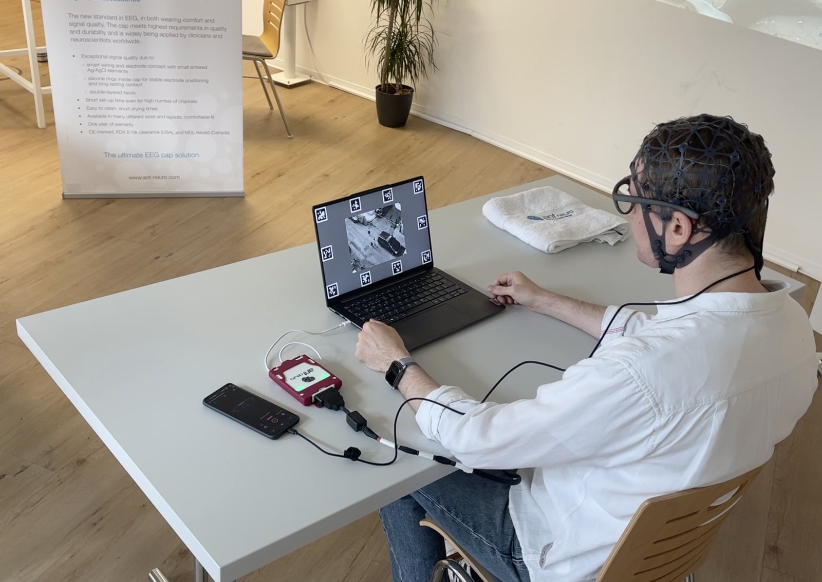 Uczestnik badania przy laptopie, w czepku EEG waveguard i okularach pomiarowych Neon, na blacie wzmacniacz eego24 i smartfon sterujący nagraniem